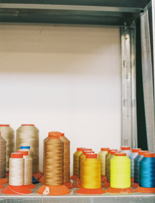 A close-up view inside a metal cabinet or shelf holding multiple spools of thread in assorted colors, including beige, brown, yellow, green, and blue, arranged on orange cone stands.