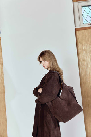Woman wearing a brown coat with a large bag against a light-colored wall.