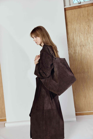 Woman wearing a long brown coat holding a matching bag indoors.