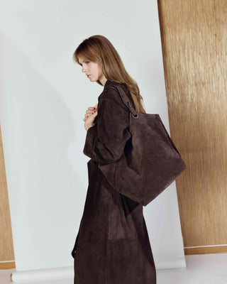 Woman wearing a long brown coat holding a matching bag indoors.