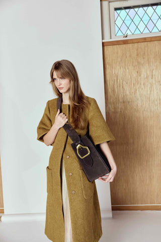 Woman wearing a long brown coat holding a brown leather bag indoors.