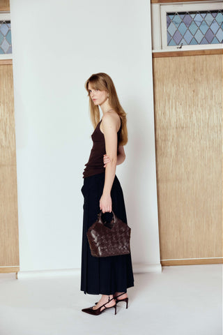 Woman holding a brown handbag in a minimalistic setting with white walls and wooden panels.