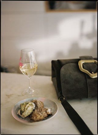 A dark brown suede shoulder bag with a gold buckle and black strap, styled on a light marble table next to a glass of white wine and a plate of pastries.